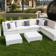 A white modular sofa with gray and white cushions sits on a green lawn in an outdoor seating area. In front are two Signature Coffee Tables by Ledge Lounger, while a modern black spherical structure stands in the background. A tall potted plant completes this elegant setting from our Signature Collection.