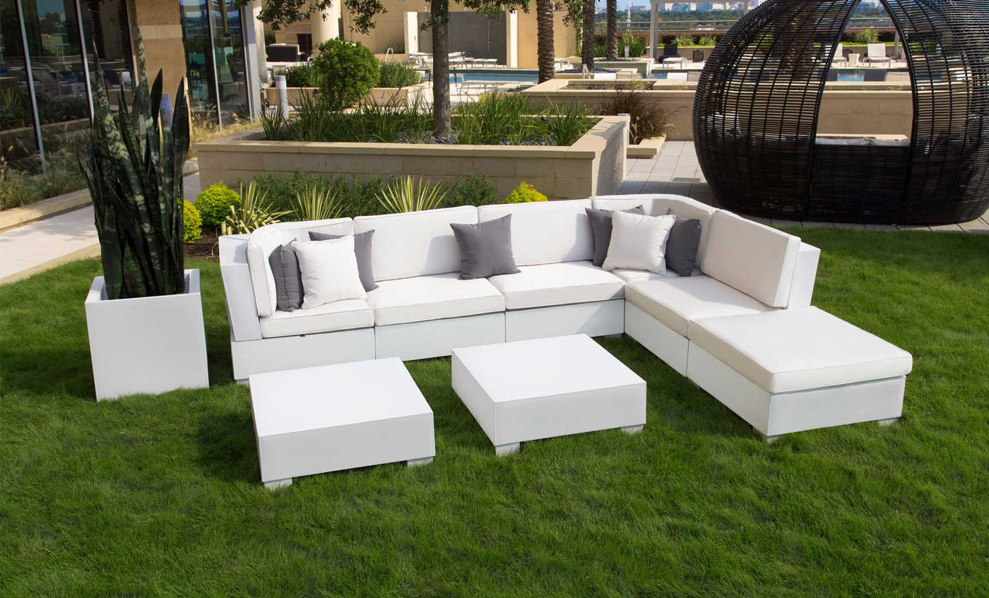 A white modular sofa with gray and white cushions sits on a green lawn in an outdoor seating area. In front are two Signature Coffee Tables by Ledge Lounger, while a modern black spherical structure stands in the background. A tall potted plant completes this elegant setting from our Signature Collection.