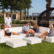 Five people converse on a white sectional sofa by Ledge Lounger on a grassy lawn. One stands with a drink, surrounded by trees, a large wicker chair, and a skyline-view building in the backdrop. A Signature Coffee Table completes the relaxing outdoor scene.