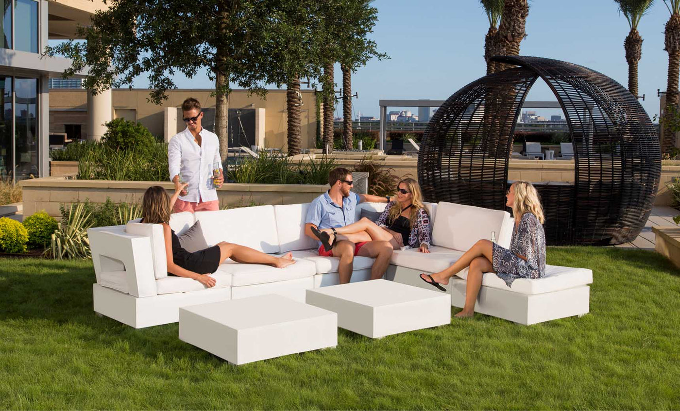 Five people converse on a white sectional sofa by Ledge Lounger on a grassy lawn. One stands with a drink, surrounded by trees, a large wicker chair, and a skyline-view building in the backdrop. A Signature Coffee Table completes the relaxing outdoor scene.