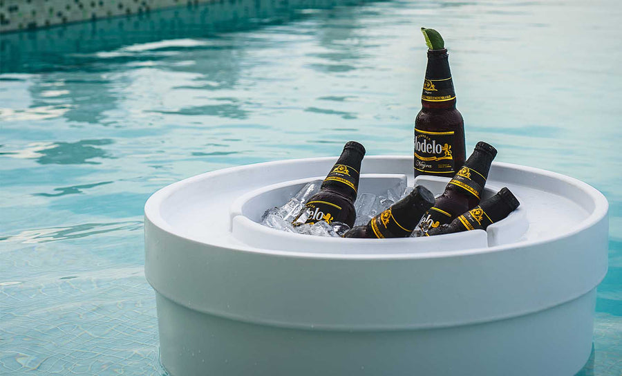Five bottles of Modelo beer chill in a Signature Ice Basin by Ledge Lounger, filled with ice and floating on a clear blue swimming pool. One bottle stands upright with a lime wedge on the rim, while sunlight reflects off the water, creating a relaxing summer ambiance.