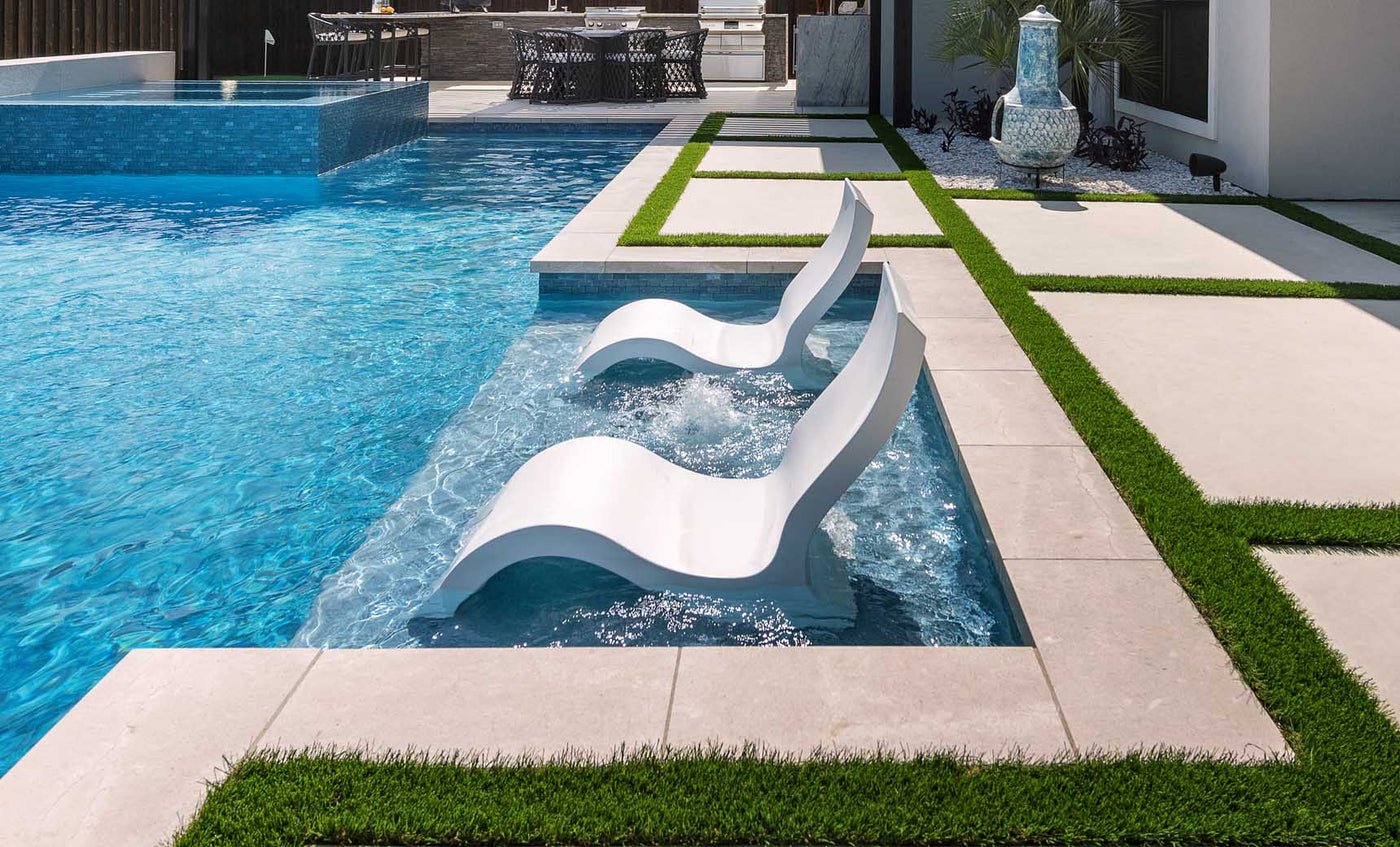 Two Ledge Lounger Signature Lowback Chairs are partially submerged in a modern pool, forming a serene outdoor oasis. A stone patio with artificial grass borders the area, while sunlight dances on the clear blue water beside a large decorative vase.