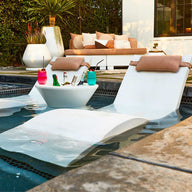 Two white loungers, partially submerged in a pool, flank a Signature Tall Ice Bin Side Table by Ledge Lounger, filled with drinks and colorful cups. A nearby patio area features a white sofa with cushions, various potted plants, and stylish Ledge Lounger Umbrellas for shade.