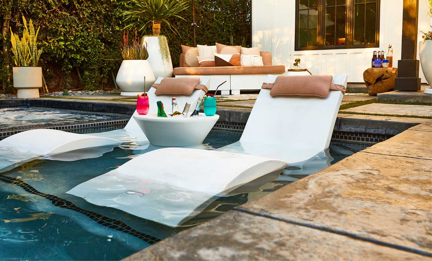 Two white loungers, partially submerged in a pool, flank a Signature Tall Ice Bin Side Table by Ledge Lounger, filled with drinks and colorful cups. A nearby patio area features a white sofa with cushions, various potted plants, and stylish Ledge Lounger Umbrellas for shade.