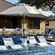 A luxurious outdoor pool area showcases white lounge chairs partially submerged in water, complemented by a sturdy umbrella base. A house with a tiled roof and cozy seating with cushions is in the background, alongside a Ledge Lounger Signature Tall Ice Bin Side Table.