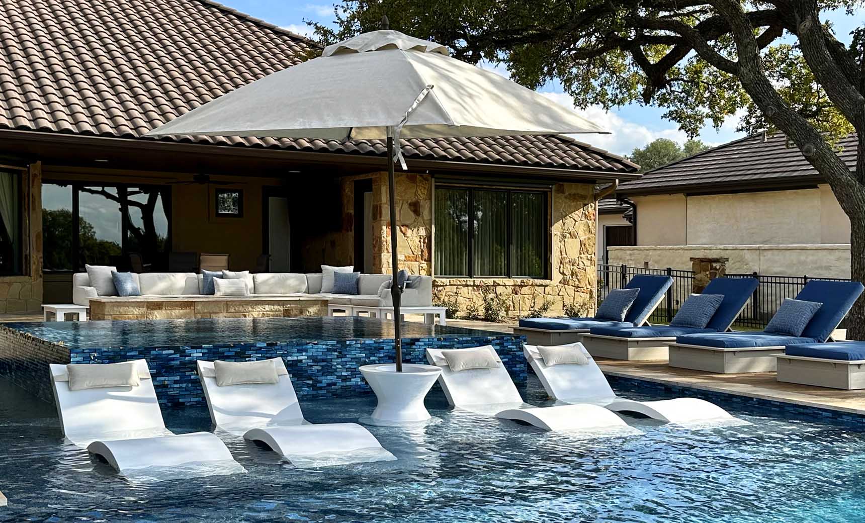 A luxurious outdoor pool area showcases white lounge chairs partially submerged in water, complemented by a sturdy umbrella base. A house with a tiled roof and cozy seating with cushions is in the background, alongside a Ledge Lounger Signature Tall Ice Bin Side Table.