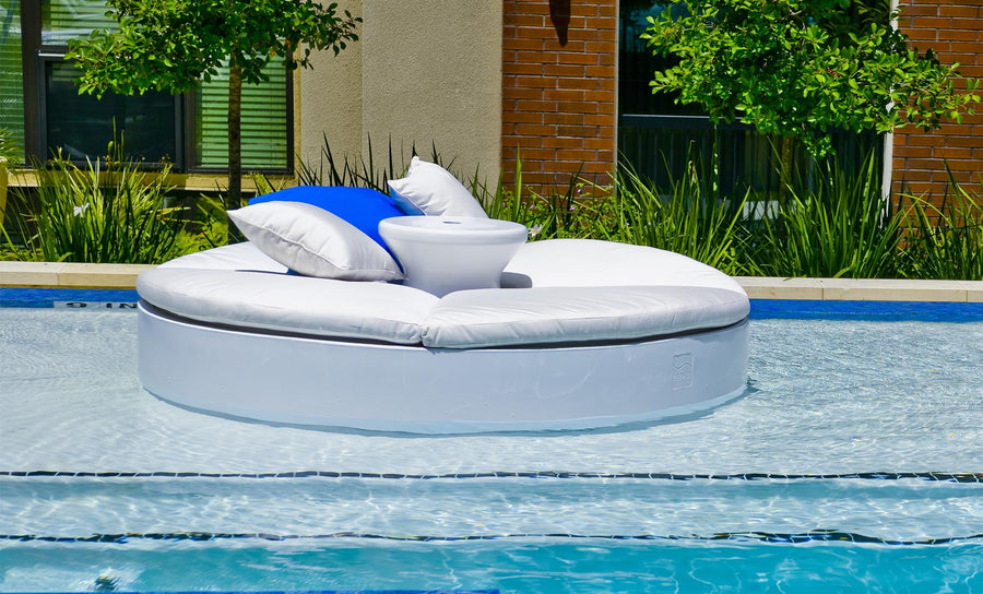 Relax in style on Ledge Loungers Signature Round Sunbed, a white circular inflatable float with cushioned seating, a central cup holder, and blue/white pillows. It sits beautifully on the water against a backdrop of bricks, lush greenery, and a nearby building.
