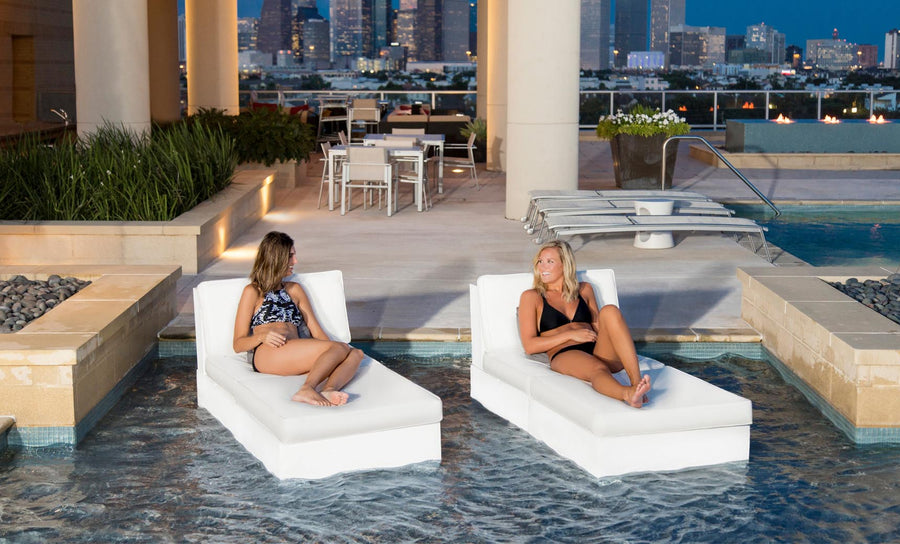 Two women relax on Ledge Loungers Signature Sectional 2 Piece Sun Chairs, partially submerged in a pool. They sit across from each other with modern architecture and a nighttime cityscape in the background, while tables and chairs on the patio complete the serene scene.