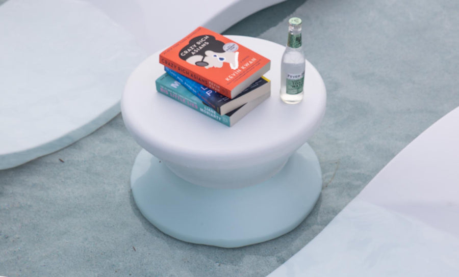 A small white Ledge Lounger Signature Standard Side Table holds three books and sparkling water, set against a textured blue and white backdrop reminiscent of waves or sand, ideal for adding elegance to your sun shelf or in-pool use.