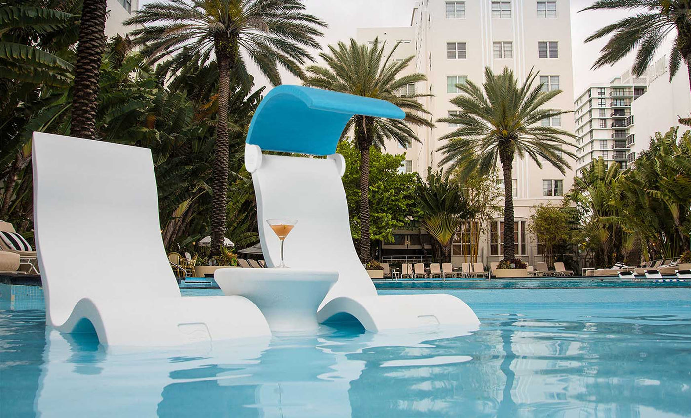 Two Signature Chairs by Ledge Lounger rest on a tanning ledge, partially submerged, with a small table holding a cocktail. Tall palm trees and a white building form the backdrop, creating an outdoor oasis with a tropical and relaxed atmosphere.