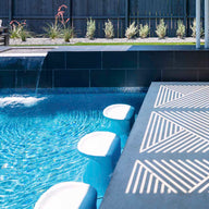 The modern pool boasts Ledge Loungers Signature Barstool Bar Height, blending seamlessly with a sleek tiled spillover edge and small waterfall. Sunlight dances on the deck as outdoor chairs and plants cozy up to a black fence—perfect for entertaining.