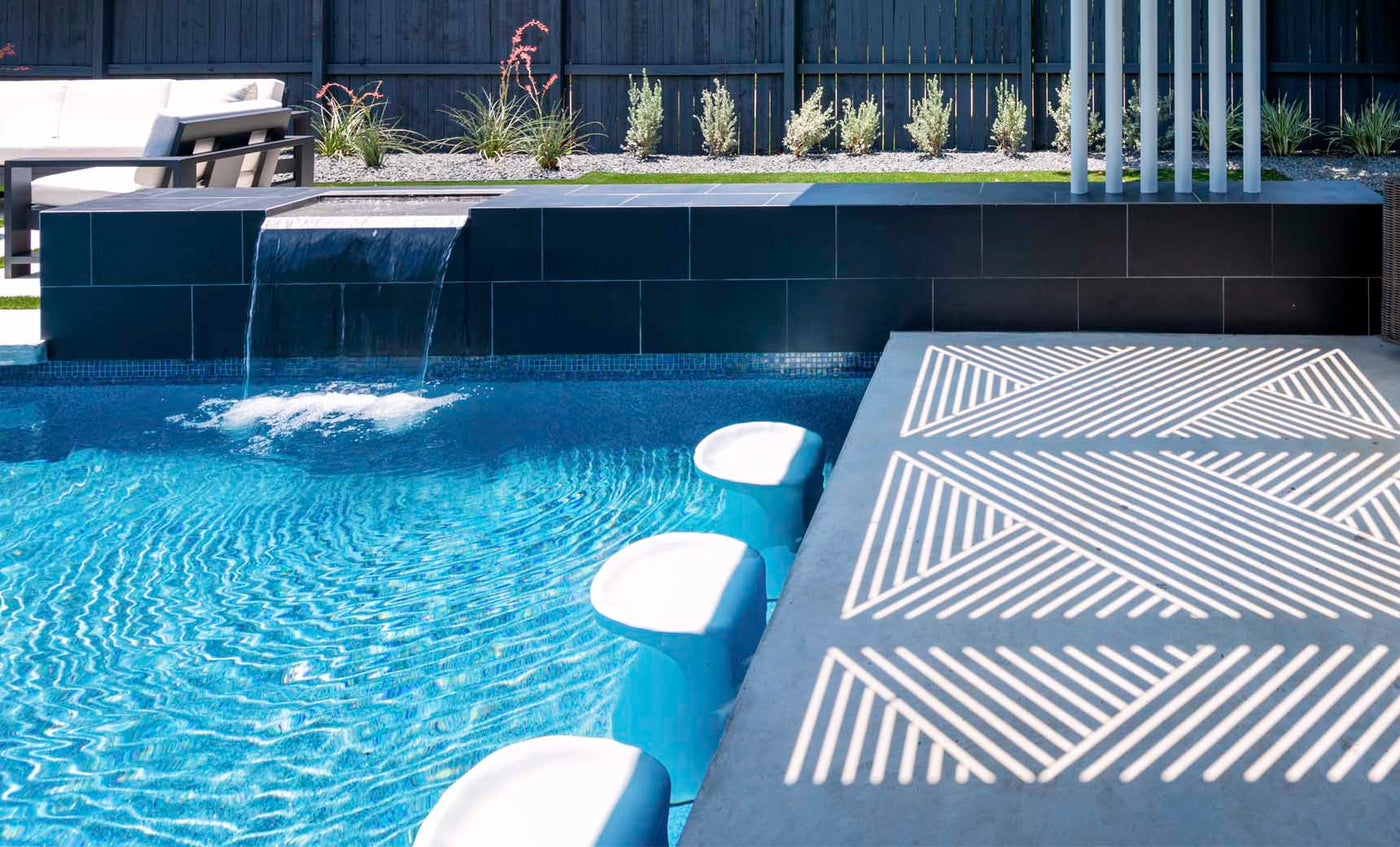 The modern pool boasts Ledge Loungers Signature Barstool Bar Height, blending seamlessly with a sleek tiled spillover edge and small waterfall. Sunlight dances on the deck as outdoor chairs and plants cozy up to a black fence—perfect for entertaining.