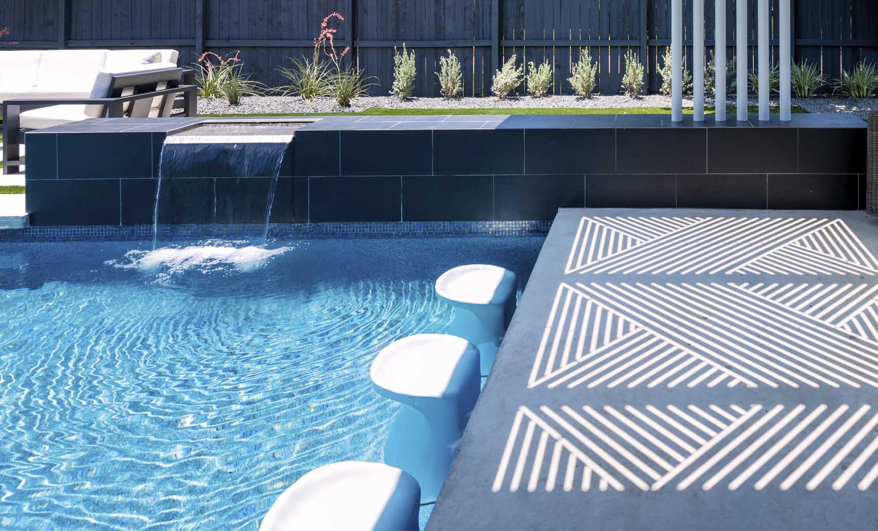 The modern pool boasts Ledge Loungers Signature Barstool Bar Height, blending seamlessly with a sleek tiled spillover edge and small waterfall. Sunlight dances on the deck as outdoor chairs and plants cozy up to a black fence—perfect for entertaining.