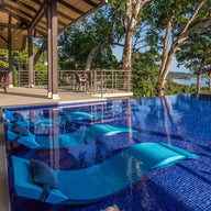 Three blue in-pool chaise loungers with matching headrest pillows on a blue tile mosaic pool ledge.
