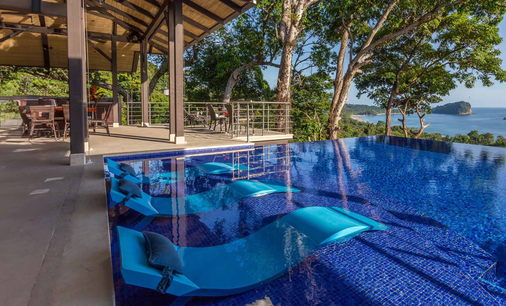 Three blue in-pool chaise loungers with matching headrest pillows on a blue tile mosaic pool ledge.