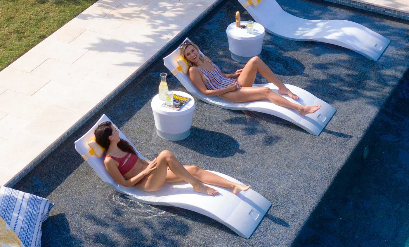 Girls smiling on in pool chaise loungers with matching headrest pillow and a side table.