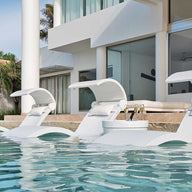 Three Ledge Lounger Signature Chaise Deep lounges rest on a tanning ledge in a pool by a modern house with large glass windows. Palm trees sway under the sunny sky, enhancing the luxurious, relaxing vibe.