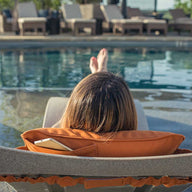 A person with long hair is lounging by the pool using a Ledge Lounger Signature Headrest Pillow on a chair with an orange cushion and a smartphone in the side pocket, while several empty chairs await others seeking customizable relaxation.