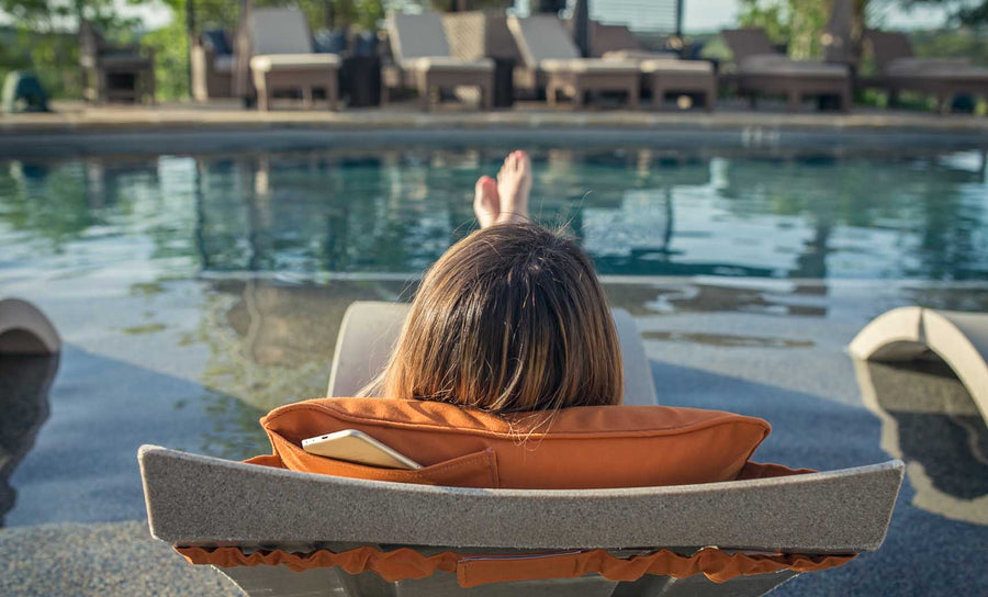 A person with long hair is lounging by the pool using a Ledge Lounger Signature Headrest Pillow on a chair with an orange cushion and a smartphone in the side pocket, while several empty chairs await others seeking customizable relaxation.