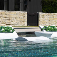 Two Ledge Lounger white chairs with green leaf towels offer in-pool relaxation, submerged in water. Between them, a round white table awaits customizable options like a refreshing drink. The setting is complemented by a stone wall and lush greenery. Signature Headrest Pillow adds extra comfort.