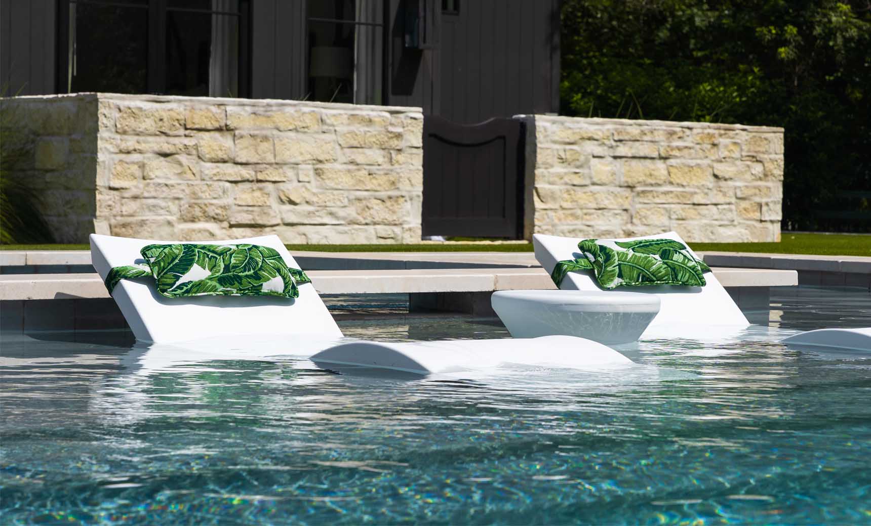 Two Ledge Lounger white chairs with green leaf towels offer in-pool relaxation, submerged in water. Between them, a round white table awaits customizable options like a refreshing drink. The setting is complemented by a stone wall and lush greenery. Signature Headrest Pillow adds extra comfort.