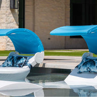 Two Ledge Lounger white loungers with blue canopies and patterned pillows, including Signature Headrest Pillows, are partially submerged in a pool near a stone building and lush grass. These loungers provide ideal in-pool relaxation with customizable options.