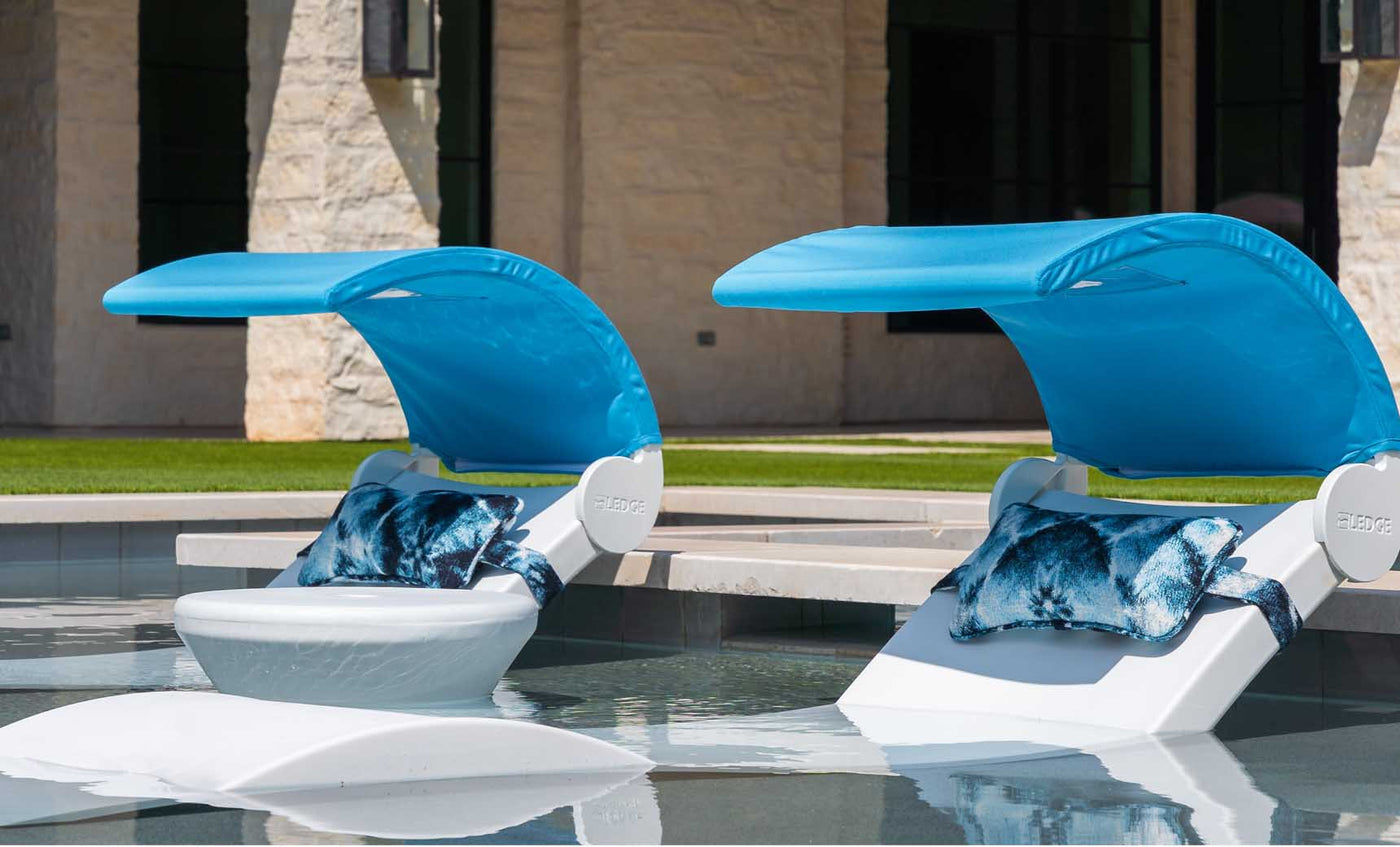 Two Ledge Lounger white loungers with blue canopies and patterned pillows, including Signature Headrest Pillows, are partially submerged in a pool near a stone building and lush grass. These loungers provide ideal in-pool relaxation with customizable options.