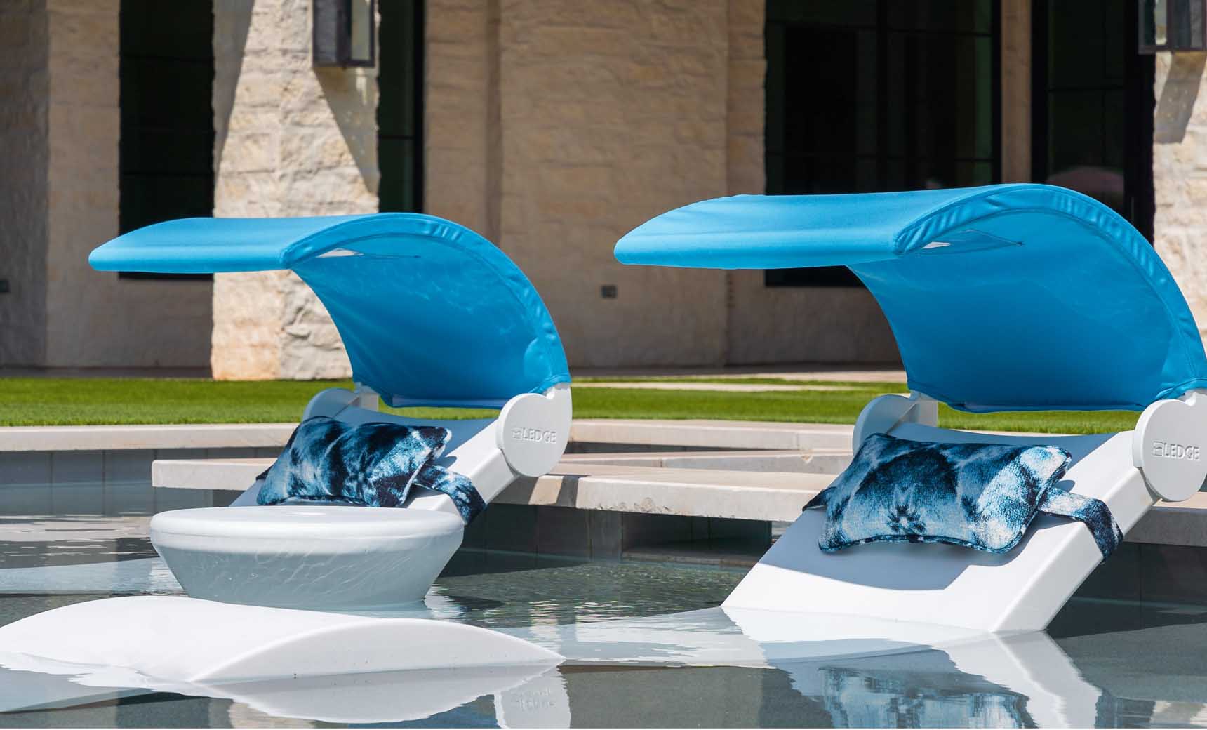 Two Ledge Lounger white loungers with blue canopies and patterned pillows, including Signature Headrest Pillows, are partially submerged in a pool near a stone building and lush grass. These loungers provide ideal in-pool relaxation with customizable options.