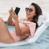 A woman in a pink swimsuit and sunglasses is laughing while holding a smartphone. She reclines on a Ledge Lounger with the Signature Headrest Pillow, partially submerged for in-pool relaxation, looking joyful and relaxed.