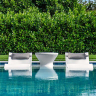Two white Ledge Lounger Signature Headrest Pillow loungers with gray cushions rest partly submerged in a clear blue pool, flanking a small white table. Lush green foliage forms a dense background, enhancing the natural tranquility of this in-pool relaxation setting.