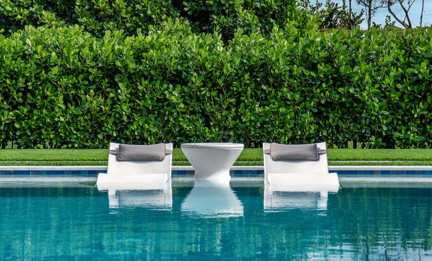 Two white Ledge Lounger Signature Headrest Pillow loungers with gray cushions rest partly submerged in a clear blue pool, flanking a small white table. Lush green foliage forms a dense background, enhancing the natural tranquility of this in-pool relaxation setting.