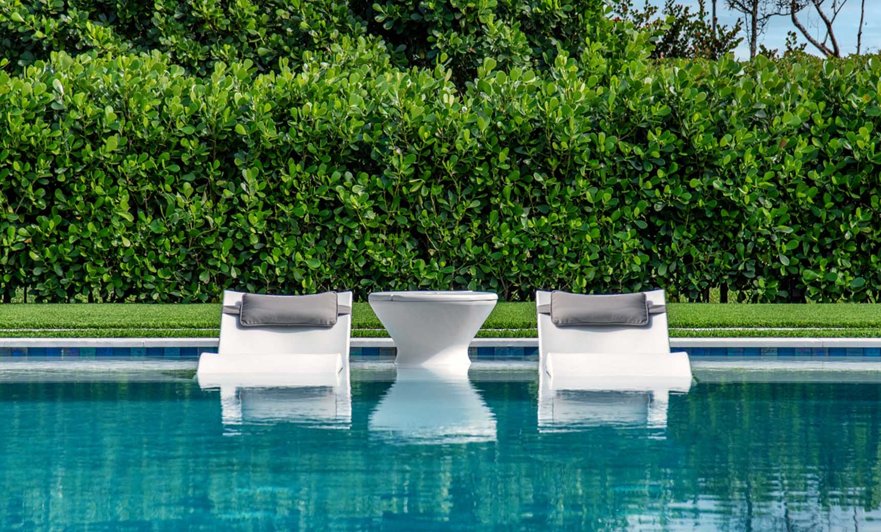 Two white Ledge Lounger Signature Headrest Pillow loungers with gray cushions rest partly submerged in a clear blue pool, flanking a small white table. Lush green foliage forms a dense background, enhancing the natural tranquility of this in-pool relaxation setting.