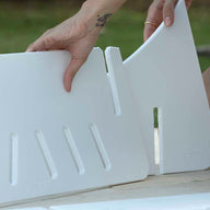 On a bright day, someone assembles white, triangular plastic Signature Chaise Deep Riser pieces with rectangular slots and directional indicators. The Ledge Lounger product features size markings, and you might notice a small tattoo on their left hand while they work.