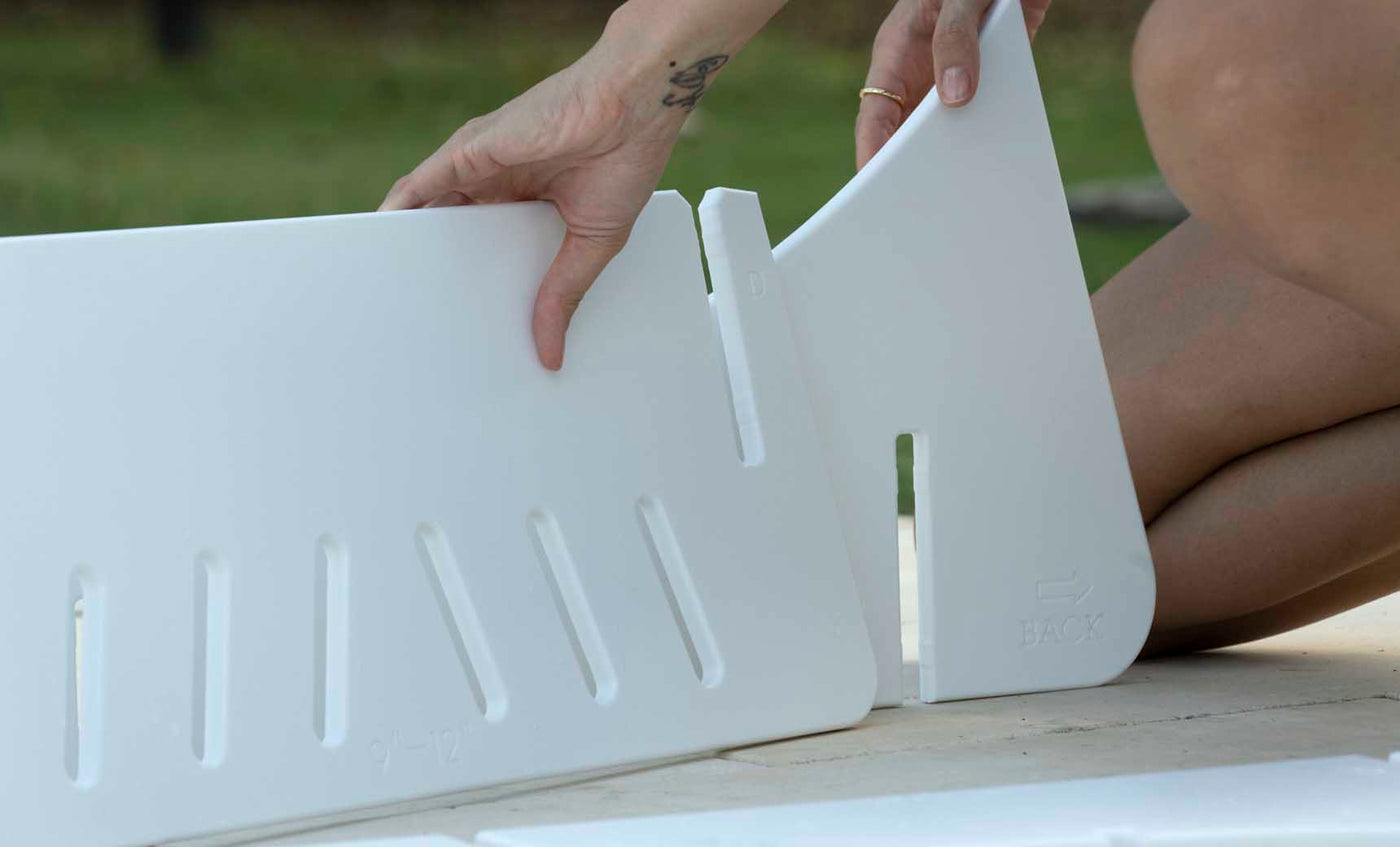 On a bright day, someone assembles white, triangular plastic Signature Chaise Deep Riser pieces with rectangular slots and directional indicators. The Ledge Lounger product features size markings, and you might notice a small tattoo on their left hand while they work.