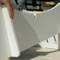 Close-up of hands assembling a white Signature Chaise Deep Riser by Ledge Lounger on a table, inserting a panel into a slot. Additional pieces and shadows are visible, evoking the crafting of outdoor day essentials.