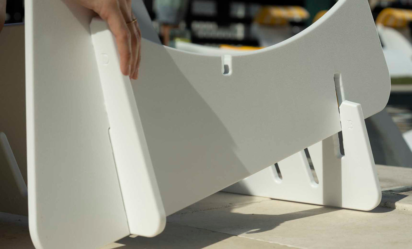 Close-up of hands assembling a white Signature Chaise Deep Riser by Ledge Lounger on a table, inserting a panel into a slot. Additional pieces and shadows are visible, evoking the crafting of outdoor day essentials.