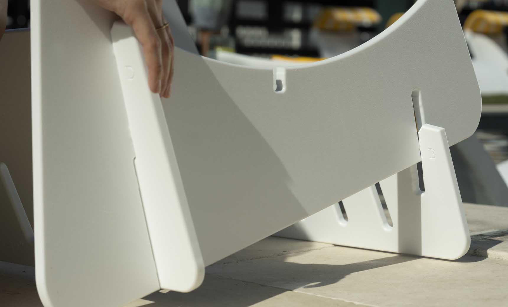 Close-up of hands assembling a white Signature Chaise Deep Riser by Ledge Lounger on a table, inserting a panel into a slot. Additional pieces and shadows are visible, evoking the crafting of outdoor day essentials.