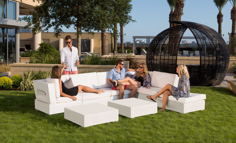 Five people relax on a Ledge Lounger Signature Sectional Corner, a modern white outdoor sofa set on grass. A man pours drinks while four others chat, with palm trees and a large circular wicker structure in the backdrop against an urban skyline.