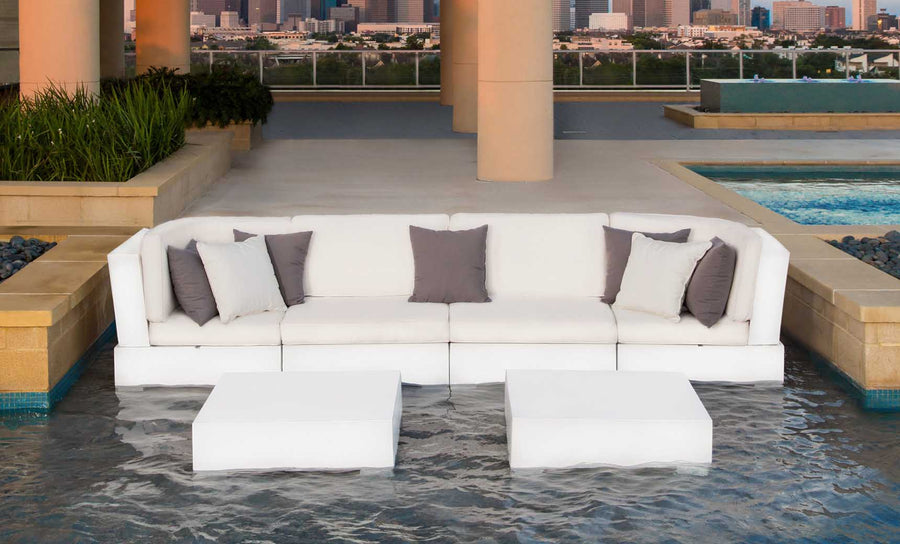 The Signature Sectional Middle by Ledge Lounger, featuring gray and white two-piece cushions, rests on shallow water with two matching low tables. The city skyline and pool view in the background enhance the modern luxury ambiance.