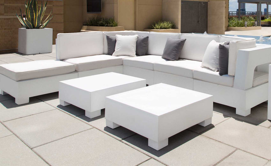The outdoor seating area boasts Ledge Loungers Signature Sectional Endcap, a white sectional sofa with gray and white cushions, and two white square tables on a tiled patio. Surrounded by potted plants and part of a water feature, it creates cozy relaxation corners.