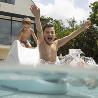 Two kids enjoy the Ledge Lounger Signature Slide, with one sliding down joyfully into a pool and another at the top. A house and trees are visible in the clear sky background.
