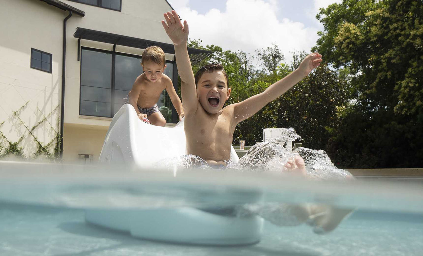 Two kids enjoy the Ledge Lounger Signature Slide, with one sliding down joyfully into a pool and another at the top. A house and trees are visible in the clear sky background.