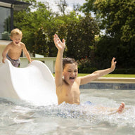 Two young boys enjoy a sunny day by the pool. One joyfully glides down the Signature Slide by Ledge Lounger with arms raised, while the other stands atop, smiling. They bask in excitement near a modern house nestled in a garden.