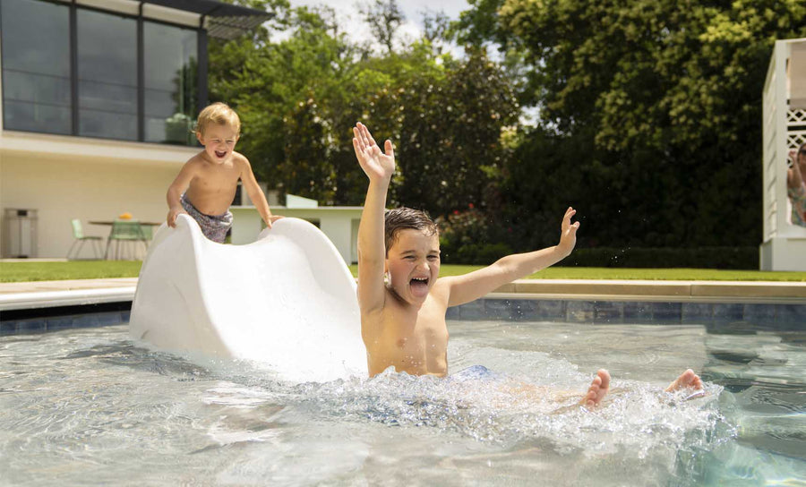 Two young boys enjoy a sunny day by the pool. One joyfully glides down the Signature Slide by Ledge Lounger with arms raised, while the other stands atop, smiling. They bask in excitement near a modern house nestled in a garden.