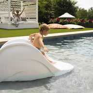 A child slides joyfully down the Ledge Lounger Signature Slide into the pool as an adult swims nearby. Two people relax in a gazebo amidst lush greenery, with umbrellas and plants creating a serene backdrop on this warm, sunny day.