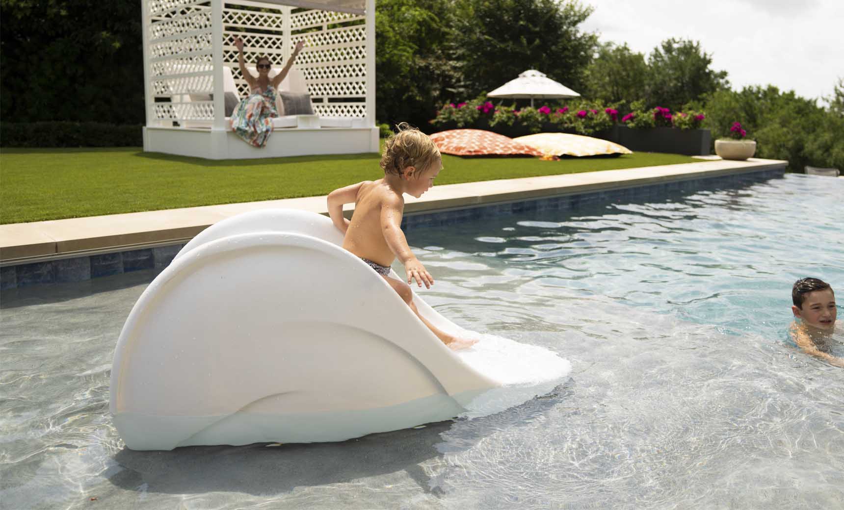 A child slides joyfully down the Ledge Lounger Signature Slide into the pool as an adult swims nearby. Two people relax in a gazebo amidst lush greenery, with umbrellas and plants creating a serene backdrop on this warm, sunny day.