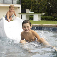 Two kids in swimsuits have fun on the Ledge Lounger Signature Slide, splashing into the pool and smiling. A woman in a floral dress and sunglasses relaxes by the tanning ledge, watching them. A building and greenery create a peaceful backdrop.