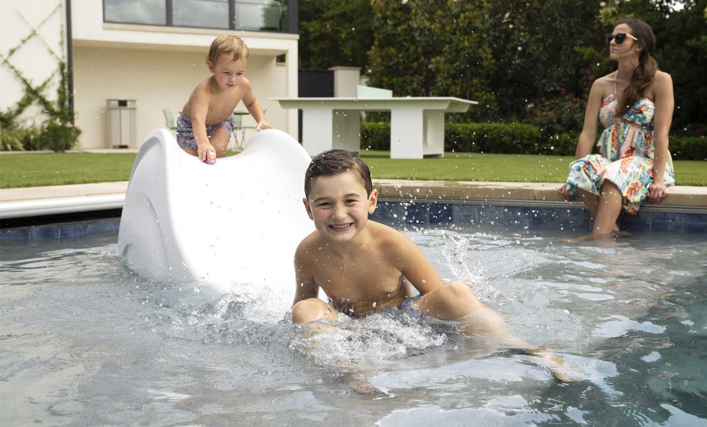 Two kids in swimsuits have fun on the Ledge Lounger Signature Slide, splashing into the pool and smiling. A woman in a floral dress and sunglasses relaxes by the tanning ledge, watching them. A building and greenery create a peaceful backdrop.