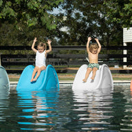 Four children joyfully sit on Ledge Loungers Signature Slide floats in a pool, hands raised. The blue and white slides shine brightly, framed by trees and a wooden fence—an idyllic summer scene on a tanning ledge.