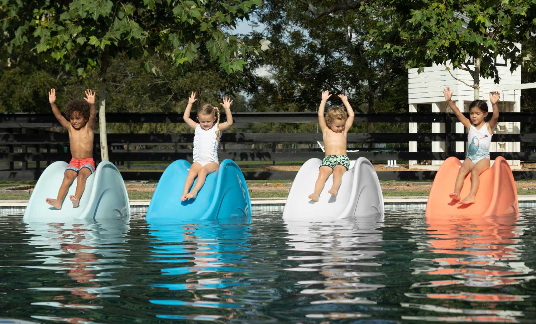 Four children joyfully sit on Ledge Loungers Signature Slide floats in a pool, hands raised. The blue and white slides shine brightly, framed by trees and a wooden fence—an idyllic summer scene on a tanning ledge.